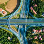 Vista aerea fotorealistica di una complessa rete stradale urbana e rurale, parzialmente coperta da ombre e vegetazione, con alcune strade chiaramente delineate come se fossero state identificate da un'IA. Obiettivo grandangolare 15mm, messa a fuoco nitida, per catturare l'estensione e la complessità del paesaggio.