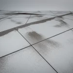 Fotografia paesaggistica grandangolare, obiettivo 15mm, che cattura un terreno geometrico astratto sotto un cielo soffuso, con vasti piani quasi piatti di pietra levigata che si intersecano ad angoli acuti ma leggermente irregolari, macchie texturizzate simili a sabbia fine sparse sui piani che rappresentano una misura, lunga esposizione che crea gradienti di luce uniformi, messa a fuoco nitida su tutta la scena.