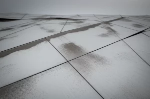 Fotografia paesaggistica grandangolare, obiettivo 15mm, che cattura un terreno geometrico astratto sotto un cielo soffuso, con vasti piani quasi piatti di pietra levigata che si intersecano ad angoli acuti ma leggermente irregolari, macchie texturizzate simili a sabbia fine sparse sui piani che rappresentano una misura, lunga esposizione che crea gradienti di luce uniformi, messa a fuoco nitida su tutta la scena.