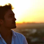 Giovane adulto che guarda con speranza verso un orizzonte luminoso dopo un periodo buio, simboleggiando la resilienza e il recupero della salute mentale. Fotografia ritratto, obiettivo da 50mm, luce calda del tramonto che illumina il volto, profondità di campo per mettere a fuoco il soggetto e sfocare lo sfondo.