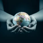 Immagine simbolica che mostra mani diverse che si uniscono delicatamente sopra un globo terrestre stilizzato e luminoso, rappresentando l'unione globale nell'ascolto e nella comprensione delle esperienze vissute col cancro. Fotografia stile prime lens 35mm, duotone blu e grigio caldo, profondità di campo accentuata sulle mani e sul globo.