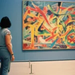 A visitor stands in a bright, modern museum gallery, looking at a large abstract painting. A subtle, dark wristband is visible on their left arm. 35mm portrait, depth of field, vibrant colors.