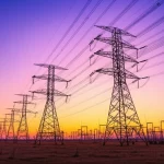 Foto ad ampio raggio di una linea di trasmissione HVDC che attraversa un paesaggio vasto, al tramonto, con i tralicci che si stagliano contro il cielo colorato, obiettivo grandangolare 10-24mm, lunga esposizione per cogliere la luce.