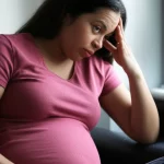 A portrait photography image, 35mm portrait, depth of field, of a pregnant woman holding her head, looking concerned, to represent the headache symptom discussed in the article.
