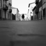 Una strada urbana deserta all'alba, vista da un'angolazione bassa. Una figura solitaria cammina in lontananza, leggermente sfocata. Obiettivo grandangolare 24mm, luce naturale soffusa, atmosfera malinconica, bianco e nero.