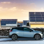 Pannelli solari fotovoltaici su tetti di un quartiere residenziale in un paesaggio innevato durante un'alba invernale, con un'auto elettrica moderna parcheggiata e collegata a una stazione di ricarica domestica. Obiettivo grandangolare, 10-24mm, lunga esposizione per nuvole soffici, messa a fuoco nitida, luce dorata dell'alba.