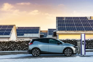 Pannelli solari fotovoltaici su tetti di un quartiere residenziale in un paesaggio innevato durante un'alba invernale, con un'auto elettrica moderna parcheggiata e collegata a una stazione di ricarica domestica. Obiettivo grandangolare, 10-24mm, lunga esposizione per nuvole soffici, messa a fuoco nitida, luce dorata dell'alba.