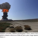 Sito di test per esplosioni controllate, obiettivo grandangolare 15mm, messa a fuoco nitida su un pennacchio di esplosione distante contro un cielo sereno, bunker di sicurezza visibili in primo piano, lunga esposizione che cattura il mosso della polvere.