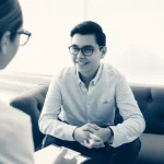Un giovane adolescente sorridente con occhiali, impegnato in una sessione di terapia individuale con un terapeuta attento in un ambiente calmo e confortevole. L'immagine trasmette speranza e supporto. Fotografia ritratto, obiettivo 35mm, duotone blu e grigio, profondità di campo.