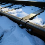 Macro fotografia, obiettivo 100mm, di un binario ferroviario in acciaio con una crepa evidente che lo attraversa. La scena è ambientata in un contesto invernale, con tracce di neve e ghiaccio sul binario e sulle traversine circostanti. L'illuminazione è fredda e laterale, per esaltare la texture metallica e la fragilità indotta dal gelo, alta definizione, messa a fuoco precisa sulla frattura del metallo.