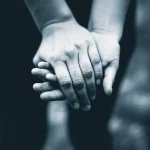 Immagine evocativa che rappresenta l'impatto emotivo della malattia di Batten su una famiglia: un primo piano di mani intrecciate, simbolo di unione e difficoltà, con uno sfondo leggermente sfocato che suggerisce l'incertezza del futuro. Prime lens, 35mm, depth of field, duotone blu e grigio per accentuare la serietà e la resilienza.