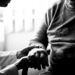 Un medico o un infermiere che tiene delicatamente la mano di un paziente anziano in un ambiente di cure palliative, trasmettendo conforto e supporto. Prime lens, 35mm, black and white film, depth of field, forte impatto emotivo.