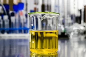 Fotografia realistica di un beaker in un laboratorio chimico contenente un liquido giallo-arancio (simulante HLLW) sopra il quale galleggia uno strato di liquido organico trasparente (D3DBDGA/dodecano). Sullo sfondo, strumentazione da laboratorio sfocata. Illuminazione da laboratorio, obiettivo prime 50mm, profondità di campo ridotta per focalizzare sul beaker.