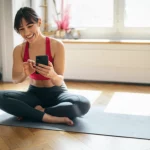 Una persona sorridente che consulta una fitness app sul suo smartphone mentre fa stretching su un tappetino in un luminoso soggiorno. Luce naturale dalla finestra. Obiettivo prime, 50mm, profondità di campo, focus sull'interfaccia dell'app.
