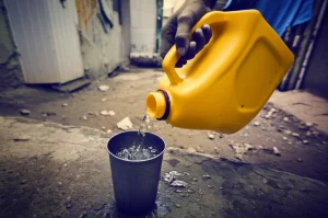 Fotografia realistica scattata con obiettivo prime 35mm che mostra una mano che versa acqua da una tanica di plastica gialla in un bicchiere metallico, in un contesto domestico umile di un insediamento informale. L'acqua appare leggermente torbida. Profondità di campo ridotta per focalizzare sull'azione. Toni colore seppia e blu duotone per un effetto suggestivo.