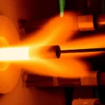 Immagine fotorealistica di un campione di lega metallica Ni-Cr-Al incandescente all'interno di un forno tubolare da laboratorio, con gas che fluisce visibilmente attorno ad esso, simulando un test di ossidazione ad alta temperatura. Obiettivo macro 70mm, illuminazione controllata per evidenziare il calore e la reazione, alta definizione dei dettagli sulla superficie della lega.