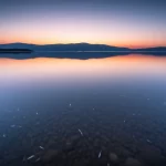 Paesaggio grandangolare, 15 mm, lago sereno che riflette il cielo, acqua in primo piano che mostra sottilmente piccole particelle di plastica, acqua liscia a lungo, focus acuto