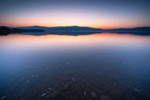 Paesaggio grandangolare, 15 mm, lago sereno che riflette il cielo, acqua in primo piano che mostra sottilmente piccole particelle di plastica, acqua liscia a lungo, focus acuto