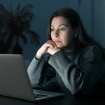 Fotografia di ritratto, primo piano di una donna di mezza età dall'aspetto stanco ma speranzoso, che guarda lo schermo di un laptop in una stanza illuminata dolcemente. Obiettivo prime 35mm, profondità di campo ridotta per sfocare lo sfondo, tonalità duotone blu e grigio per un'atmosfera calma ma riflessiva.