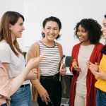 Un gruppo diversificato di donne BIPOC docenti universitarie sorridenti e impegnate in una discussione collaborativa in un'aula universitaria moderna e luminosa. Fotografia di ritratto di gruppo, obiettivo da 35mm, luce naturale, colori caldi e accoglienti, profondità di campo che enfatizza le interazioni positive.