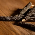 Primo piano fotorealistico di frutti di Piper longum essiccati su una superficie di legno rustico, obiettivo macro 100mm, illuminazione laterale morbida che ne esalta la texture e i dettagli, suggerendo il potenziale naturale della pianta.