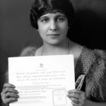 Ritratto fotografico stile anni '20 di una donna spagnola dallo sguardo intelligente e sicuro, che tiene in mano un certificato azionario. Luce laterale drammatica, bianco e nero con leggeri toni seppia, obiettivo 35mm, profondità di campo che sfoca leggermente lo sfondo di un interno borghese.