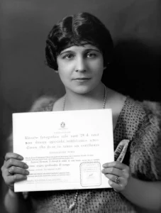 Ritratto fotografico stile anni '20 di una donna spagnola dallo sguardo intelligente e sicuro, che tiene in mano un certificato azionario. Luce laterale drammatica, bianco e nero con leggeri toni seppia, obiettivo 35mm, profondità di campo che sfoca leggermente lo sfondo di un interno borghese.
