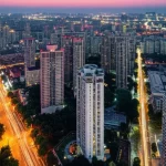 Veduta grandangolare di un quartiere urbano densamente popolato in Cina al tramonto, con un mix di edifici residenziali moderni e più vecchi, alcuni con segni di usura. Obiettivo grandangolare 20mm, lunga esposizione per catturare le luci della città, mettendo in evidenza la complessità abitativa e le diverse condizioni degli alloggi.