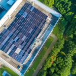 Fotografia aerea con drone di un moderno edificio universitario con un esteso impianto fotovoltaico sul tetto che combina pannelli monocristallini (neri), policristallini (bluastri) e a film sottile (neri uniformi), luce solare diretta del primo pomeriggio, alta risoluzione, dettagli nitidi dell'installazione e dell'ambiente circostante verdeggiante tipico della Malesia.