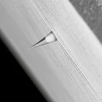 Immagine fotorealistica di una frattura che si propaga perpendicolarmente attraverso l'interfaccia tra due materiali con diverse proprietà meccaniche. La frattura è illuminata per evidenziare la sua punta. Dettaglio macro con obiettivo da 60mm, illuminazione controllata per enfatizzare le texture dei materiali e la linea di frattura.