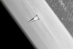 Immagine fotorealistica di una frattura che si propaga perpendicolarmente attraverso l'interfaccia tra due materiali con diverse proprietà meccaniche. La frattura è illuminata per evidenziare la sua punta. Dettaglio macro con obiettivo da 60mm, illuminazione controllata per enfatizzare le texture dei materiali e la linea di frattura.