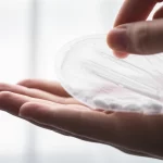 Una mano che tiene delicatamente una sacca per stomia semitrasparente, attraverso cui si intravedono alcune pillole intatte. Sfondo neutro e luminoso, focus sulla sacca e sulle pillole. Lente prime 50mm, profondità di campo ridotta per enfatizzare il soggetto.