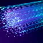 Immagine fotorealistica di un fascio di luce laser colorato (ciano e magenta duotone) che si propaga attraverso una complessa struttura cristallina o una fibra ottica, mostrando effetti di focalizzazione e dispersione non lineare, obiettivo macro 85mm, profondità di campo ridotta, illuminazione high-key per enfatizzare la luce, alta definizione.