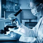 Ricercatrice in un laboratorio high-tech che osserva campioni al microscopio, con schermi che mostrano dati molecolari sullo sfondo. Illuminazione da studio controllata, obiettivo prime da 35mm, profondità di campo ridotta per mettere a fuoco la ricercatrice, toni blu e grigi duotone per un'atmosfera scientifica e speranzosa.