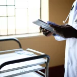 Immagine fotorealistica di un medico o infermiere in un ospedale del Kenya occidentale che esamina una cartella clinica, con blister di antibiotici visibili su un carrello vicino, luce naturale da una finestra, obiettivo 50mm, profondità di campo.