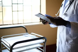 Immagine fotorealistica di un medico o infermiere in un ospedale del Kenya occidentale che esamina una cartella clinica, con blister di antibiotici visibili su un carrello vicino, luce naturale da una finestra, obiettivo 50mm, profondità di campo.
