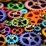 Immagine concettuale che rappresenta la complessità della misurazione dello sviluppo comunicativo in bambini autistici: una serie di ingranaggi colorati e interconnessi di diverse dimensioni, alcuni luminosi e altri più opachi, a simboleggiare le diverse misure e la loro variabile sensibilità. Obiettivo macro da 90mm, alta definizione, illuminazione da studio per enfatizzare texture e colori.