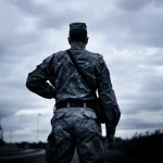 Immagine simbolica che rappresenta la solitudine e la pressione psicologica: un militare in uniforme, visto di spalle, guarda fuori da una finestra di una caserma al crepuscolo. Obiettivo prime da 50mm, toni blu e grigi duotone, profondità di campo che sfoca lo sfondo per enfatizzare l'isolamento e la riflessione interiore.