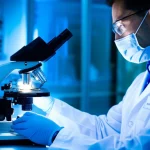 Primo piano di un ricercatore in camice bianco che osserva attentamente al microscopio un campione di linfonodo metastatico, illuminato da una luce fredda da laboratorio. L'immagine è scattata con un obiettivo macro da 100mm per enfatizzare l'alta definizione dei dettagli del campione e la concentrazione del ricercatore, simboleggiando l'ottimizzazione della terapia con carboplatino tramite LDDS.