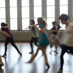 Gruppo eterogeneo di studenti universitari che partecipano con gioia a una sessione di danza espressiva in uno studio luminoso e spazioso. Obiettivo grandangolare 24mm per catturare l'ampiezza della scena e l'interazione del gruppo, luce naturale diffusa che entra da grandi finestre, messa a fuoco nitida sul gruppo ma con leggero motion blur per suggerire il movimento. Atmosfera energica e positiva.