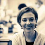 Ritratto di una donna matura e sorridente, sui 55 anni, che simboleggia la salute in menopausa, con uno sfondo leggermente sfocato di un laboratorio di ricerca. Fotografia ritratto, obiettivo 35mm, duotone seppia e blu, profondità di campo.