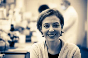 Ritratto di una donna matura e sorridente, sui 55 anni, che simboleggia la salute in menopausa, con uno sfondo leggermente sfocato di un laboratorio di ricerca. Fotografia ritratto, obiettivo 35mm, duotone seppia e blu, profondità di campo.