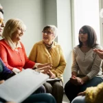Fotografia realistica di un gruppo eterogeneo di persone anziane, diverse etnie e background, che partecipano attivamente a una discussione sulla salute comunitaria in un ambiente accogliente e luminoso. Alcuni prendono appunti, altri parlano gesticolando. Si percepisce un senso di collaborazione e ascolto reciproco. Obiettivo prime 35mm, profondità di campo che mette a fuoco il gruppo ma sfoca leggermente lo sfondo, luce naturale controllata proveniente da una finestra laterale, toni caldi.