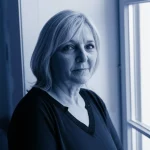 Ritratto fotografico, obiettivo 35mm, di una donna di mezza età dall'espressione pensierosa ma serena, che guarda fuori da una finestra. Luce naturale morbida, leggero effetto duotone blu e grigio, profondità di campo che sfoca leggermente lo sfondo, simboleggiando la riflessione sulla gestione a lungo termine di una condizione cronica come l'epilessia e la comparsa inaspettata di effetti collaterali.