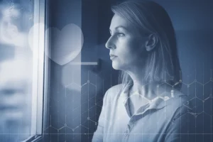Donna dall'aspetto pensieroso che guarda fuori da una finestra, simboleggiando la preoccupazione per la fertilità, con un leggero overlay grafico di un cuore e un grafico metabolico. Fotografia ritratto, 35mm, toni duotone blu e grigio, profondità di campo.