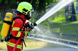 Vigile del fuoco in piena azione durante un intervento, la sua divisa tecnica innovativa ben visibile mentre maneggia una lancia antincendio, fotografia d'azione con teleobiettivo zoom 100-400mm, inseguimento del movimento, alta velocità dell'otturatore per congelare l'azione, gocce d'acqua sospese nell'aria.