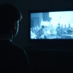 Immagine simbolica: un adolescente visto di spalle guarda uno schermo luminoso (TV o computer) che mostra immagini astratte e disturbanti di conflitto, la stanza è buia. Obiettivo 50mm, luce dello schermo come unica fonte luminosa, colori desaturati, effetto grana pellicola.
