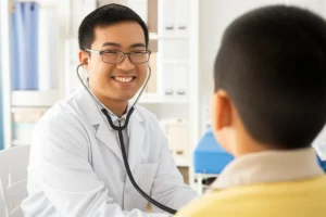 Un medico pediatra esperto, di etnia asiatica, sorridente e rassicurante, interagisce con un bambino piccolo durante una visita in un ambulatorio pediatrico luminoso e colorato. Il medico tiene uno stetoscopio. Prime lens, 35mm, depth of field, colori caldi e accoglienti.