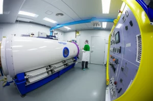 Interno di una camera iperbarica clinica moderna, vista grandangolare 18mm, illuminazione controllata, con dettagli visibili dei sistemi di monitoraggio e erogazione ossigeno, atmosfera serena e tecnologica, un tecnico osserva un pannello di controllo.