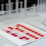 Primo piano di diverse card qDBS con campioni di sangue essiccato, disposte su un tavolo da laboratorio accanto a micropipette e provette. Illuminazione da studio, obiettivo macro 60mm, alta definizione per mostrare la texture della carta e del sangue.